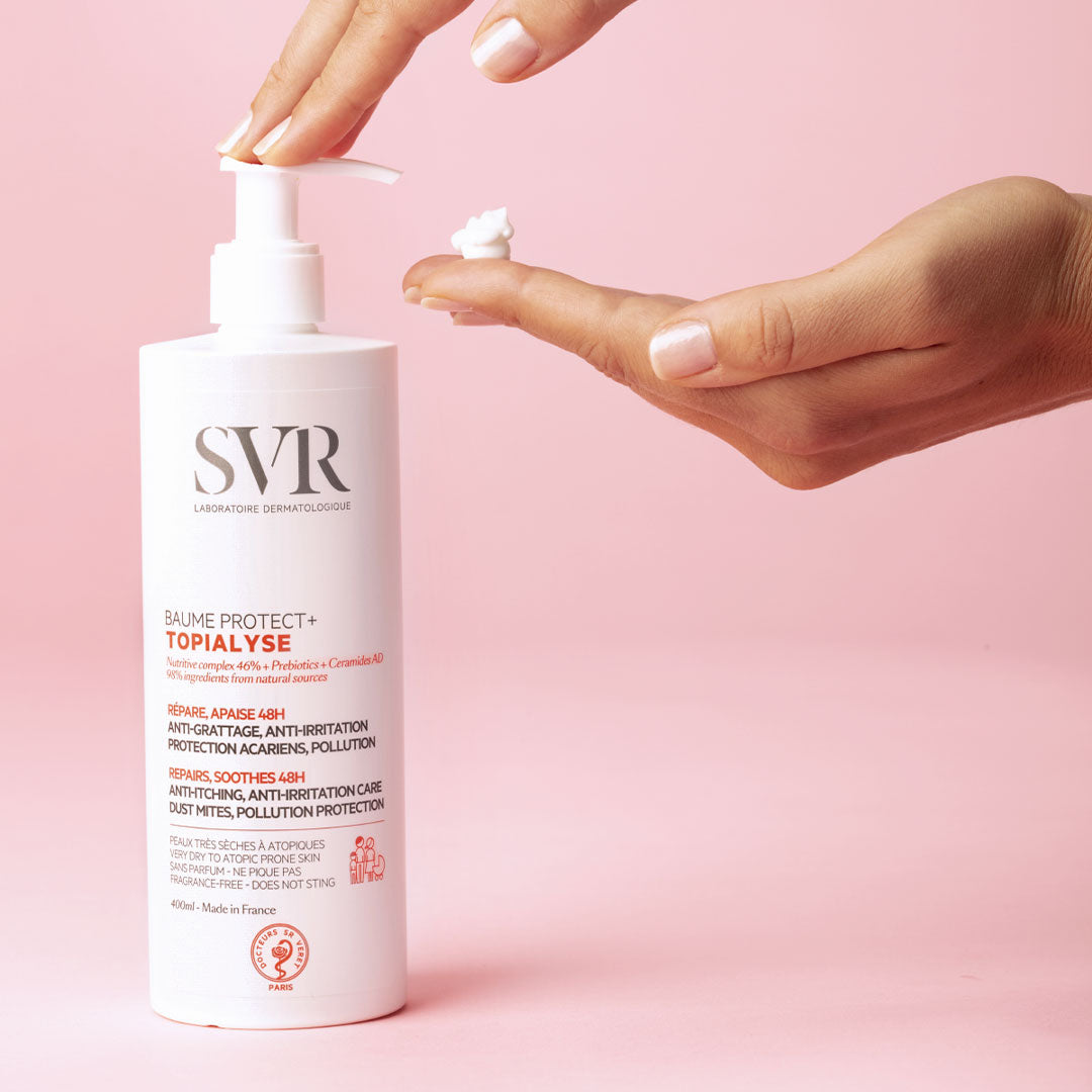 Flacon pompe blanc SVR TOPIALYSE Baume Protect+ avec une main pressant une noisette de crème sur fond rose.