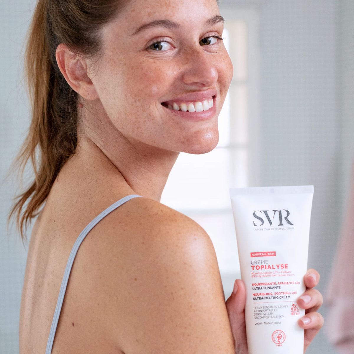 Femme souriante tenant un tube blanc de crème hydratante SVR TOPIALYSE de 200ml pour peaux sèches et sensibles.