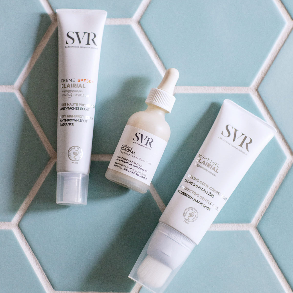 Trois produits de soin SVR Clairial sur un carrelage bleu clair : un tube de crème SPF50+, un flacon compte-gouttes et un tube.