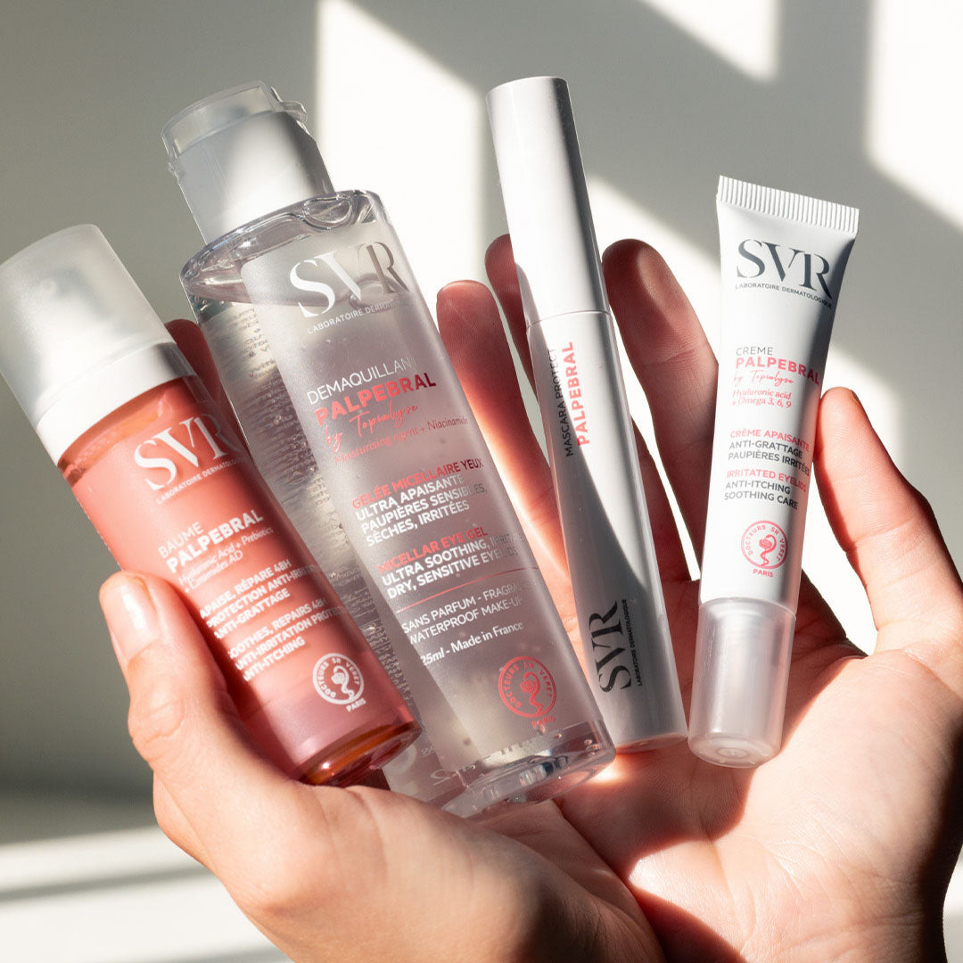 Main tenue en main présentant quatre produits SVR Palpebral : baume rose, démaquillant gel transparent, mascara et crème apaisante.
