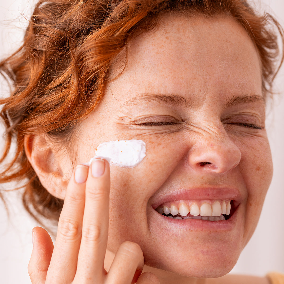 Femme rousse souriante appliquant une crème blanche sur sa joue pour revitaliser et illuminer son teint.