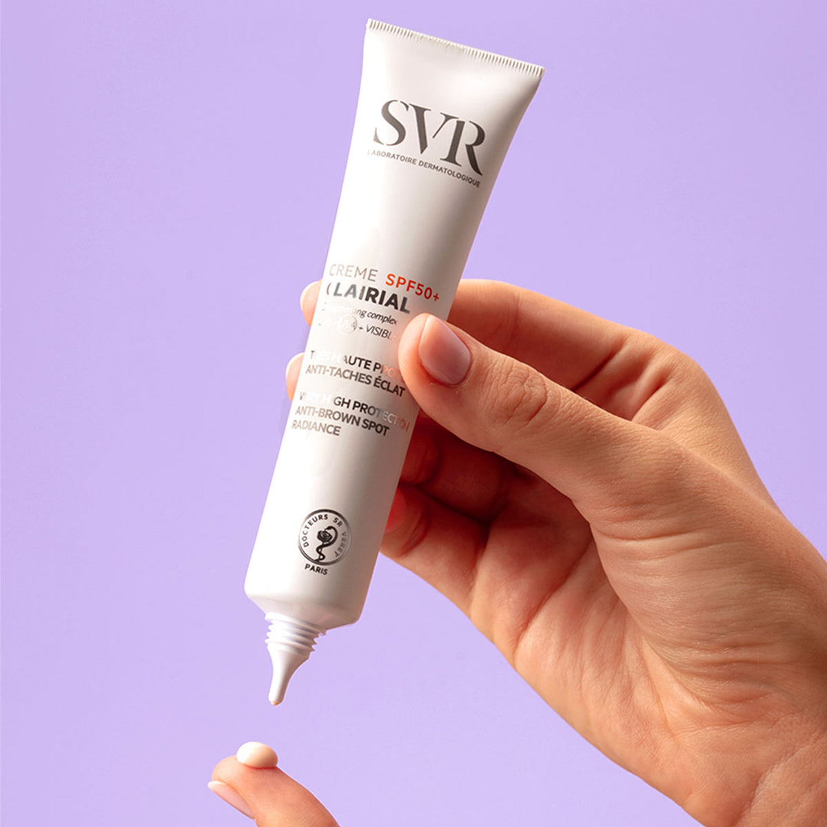 Main tenue en main pressant un tube blanc de crème solaire SVR Clairial SPF50+ sur un fond lilas.