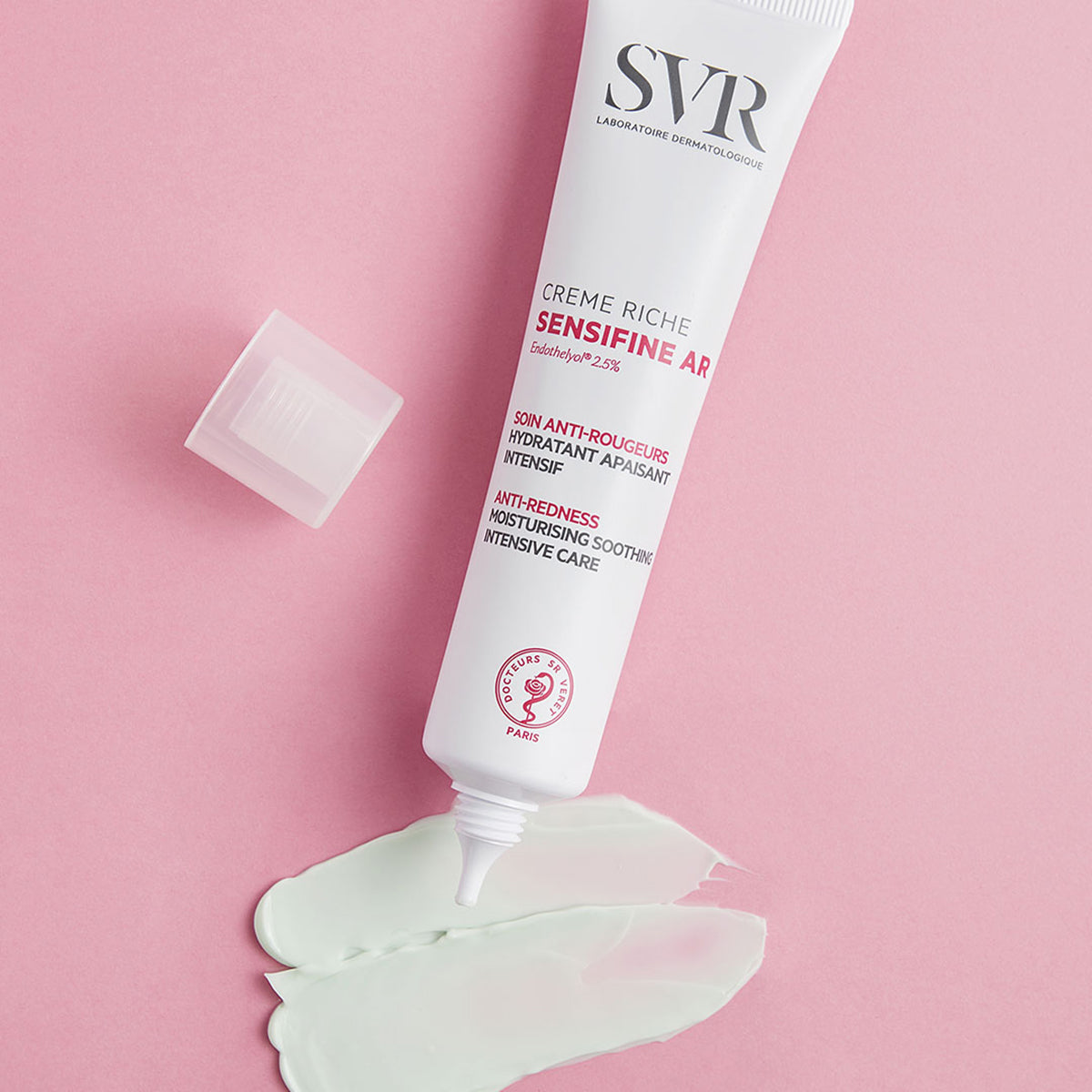 Tube blanc de crème SVR Sensifine AR avec bouchon ouvert et texture crème verte sur fond rose pastel.