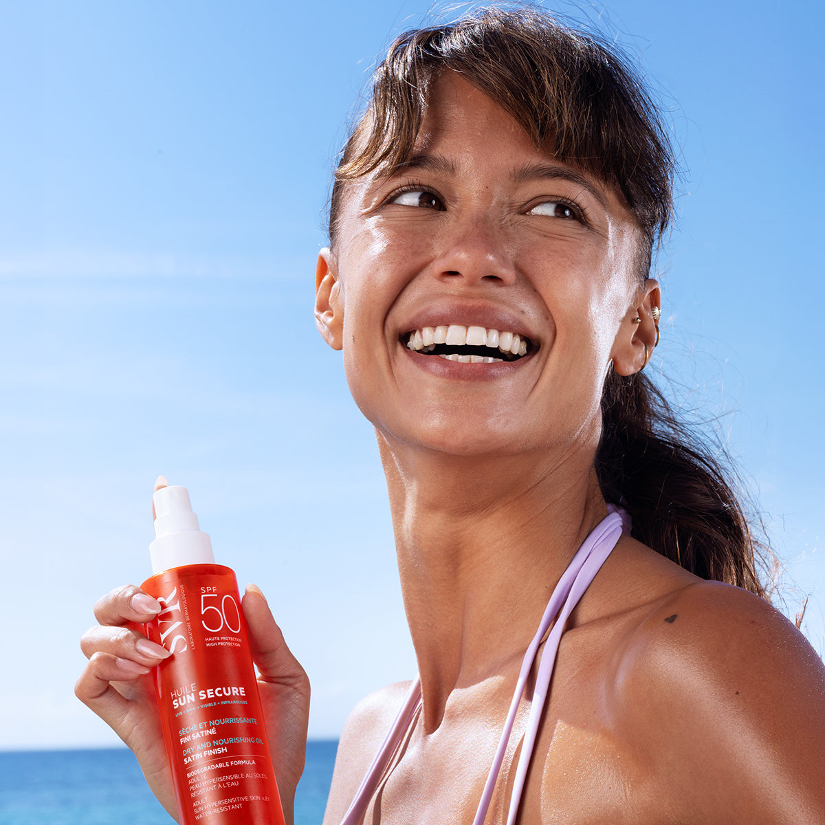 Femme souriante tenant un flacon orange de SVR Sun Secure Huile SPF50 sur fond de ciel bleu et mer ensoleillée.