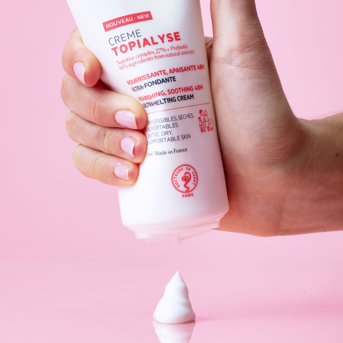 Main tenue pressant un tube blanc de crème hydratante TOPIALYSE avec une noisette de produit onctueux sur fond rose.