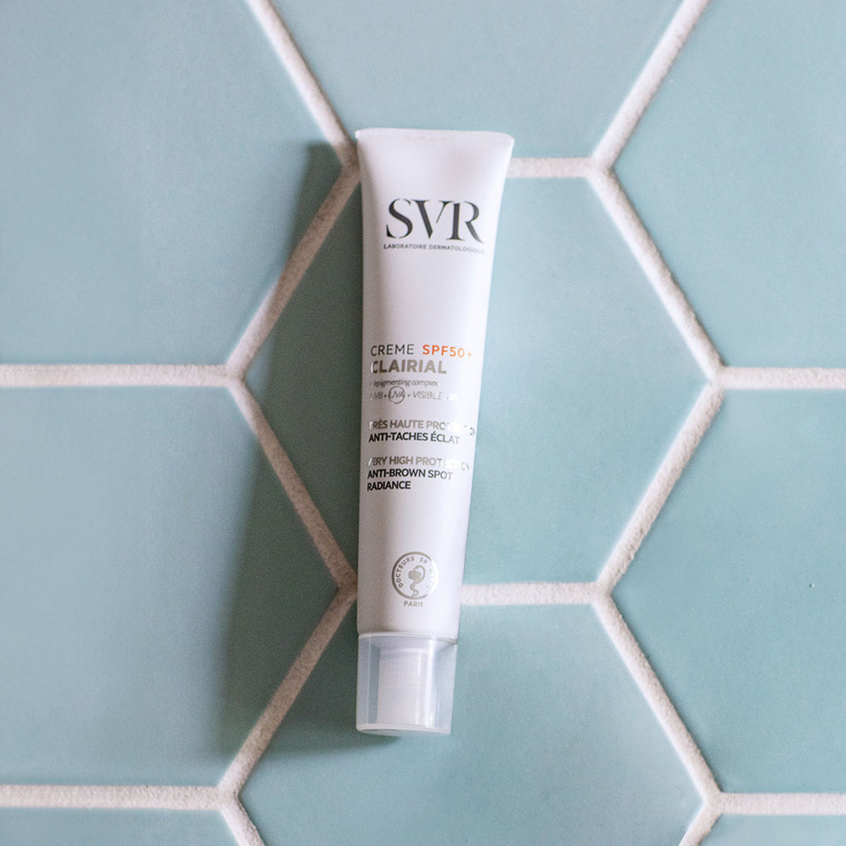 Tube blanc de crème solaire SVR Clairial SPF50+ posé sur un carrelage mural hexagonal bleu clair.