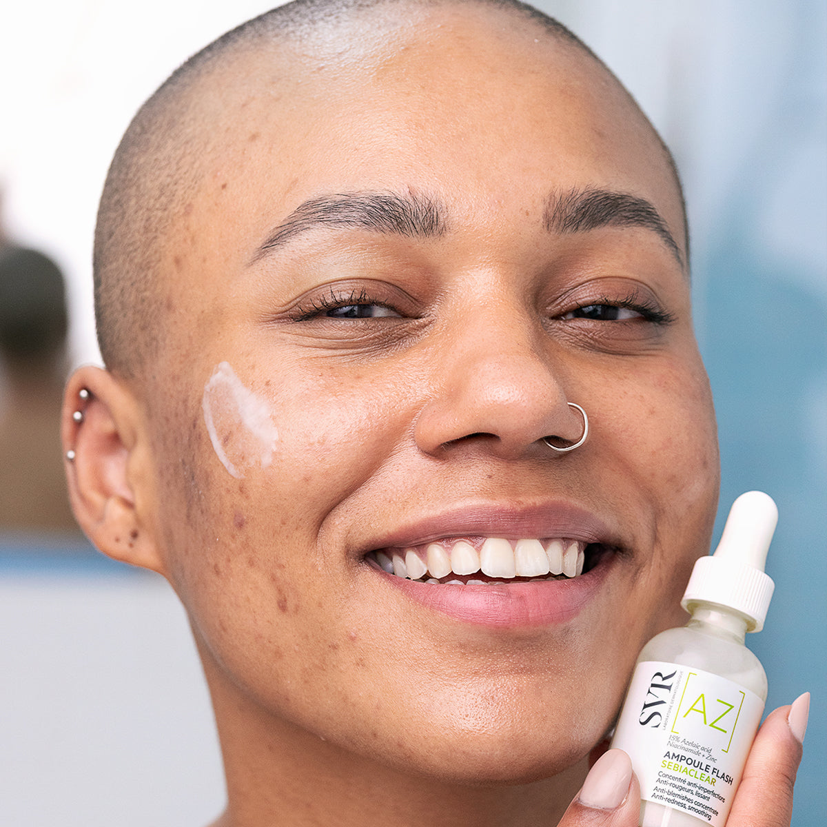 Femme souriante tenant le flacon blanc et vert du sérum SVR Sebiaclear Ampoule Flash avec une touche de crème sur la joue.