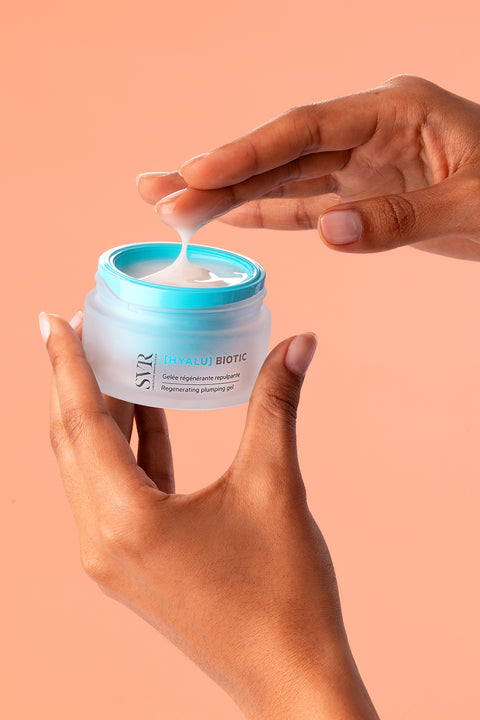 Hands holding a jar of SVR Hyalu Biotic regenerating plumping gel with a white cream texture on a peach background.