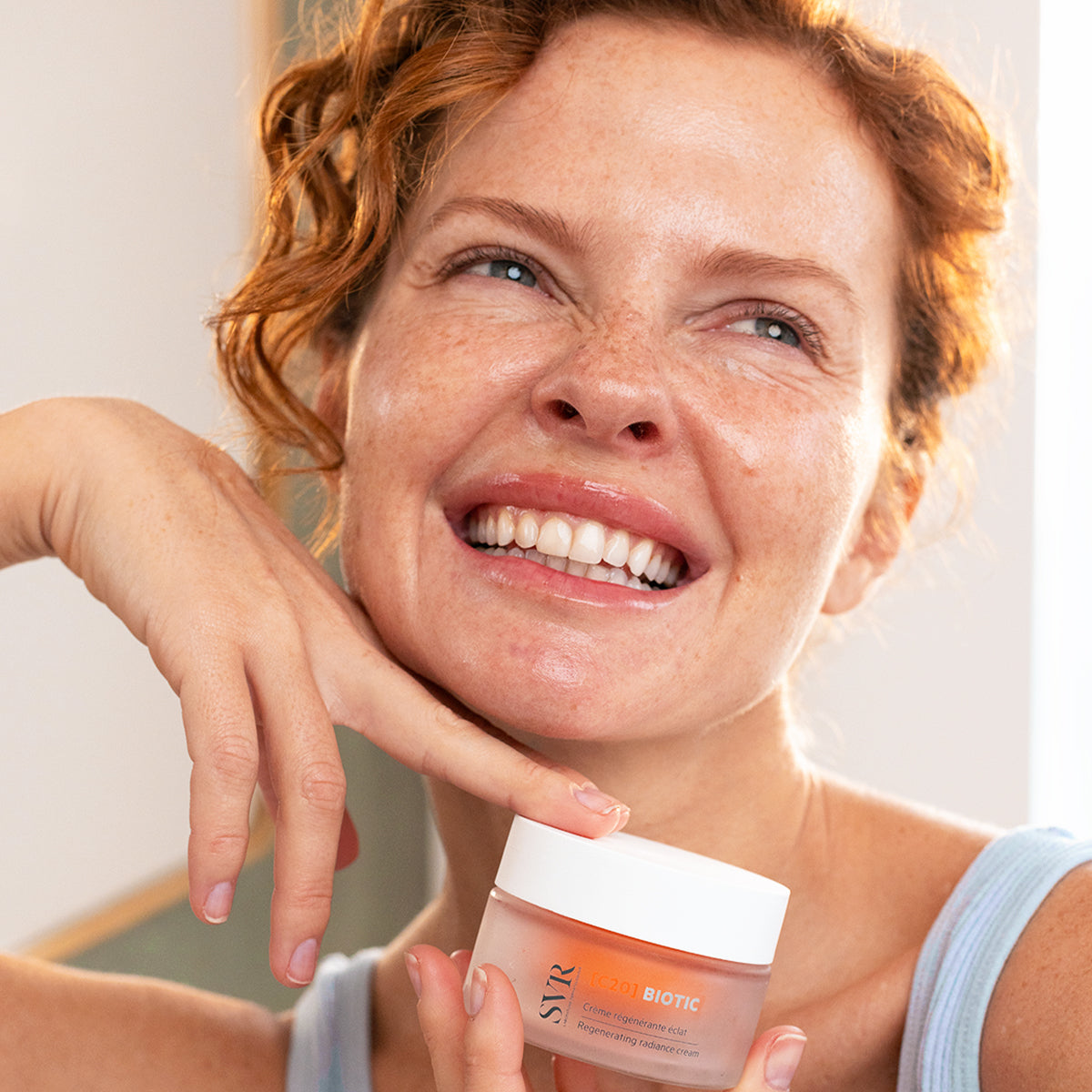 Femme souriante tenant un pot de crème régénérante SVR [C20]Biotic au design minimaliste blanc et orangé.